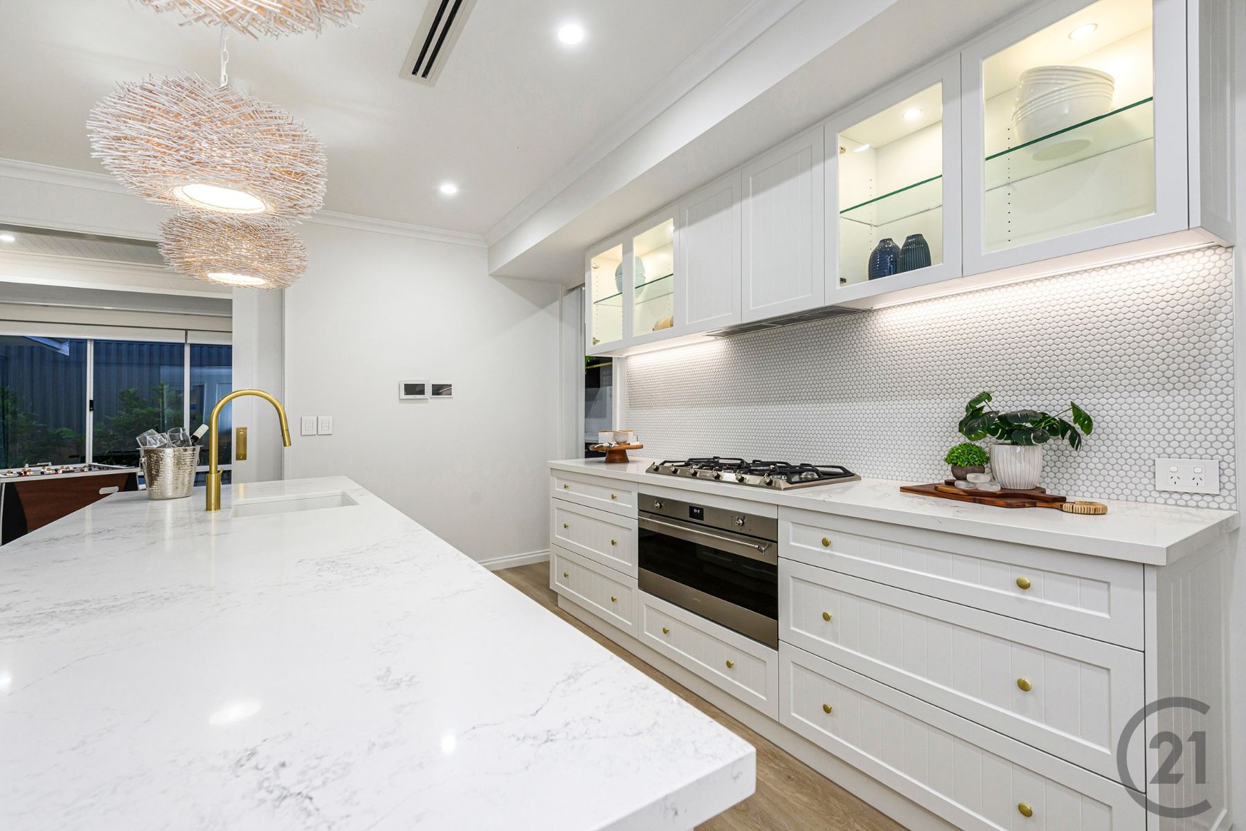 White kitchen with marble-look stone island, brass mixer, rattan pendants and lit glass cabinets